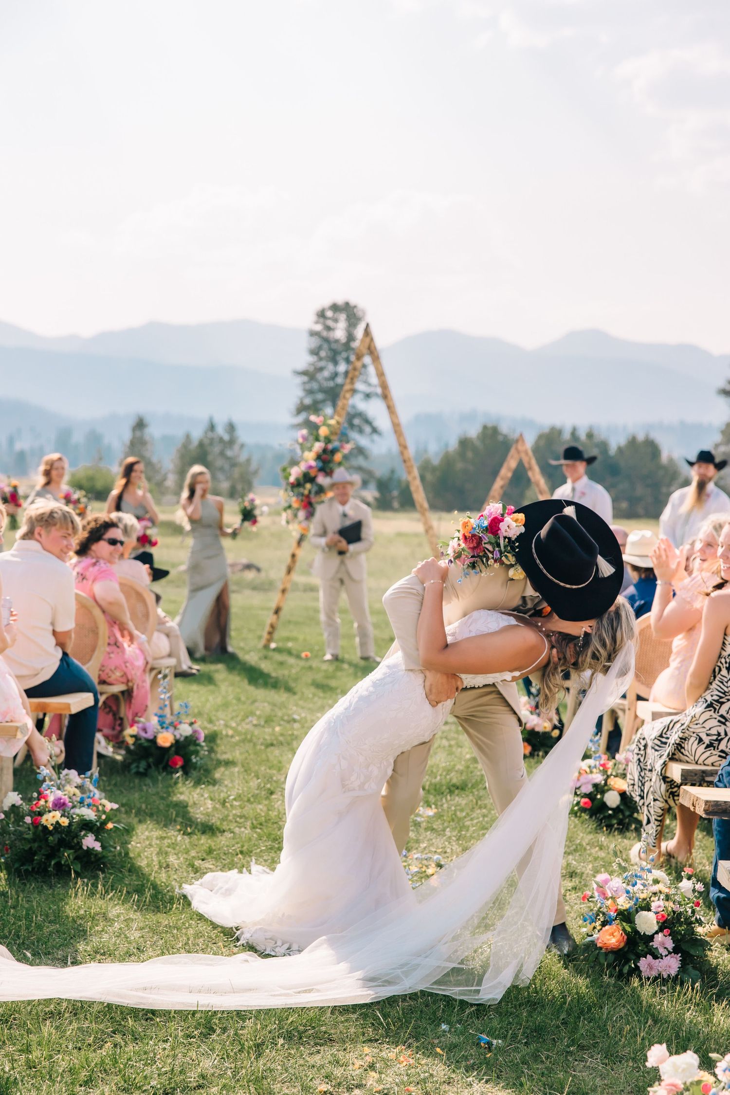 A romantic mountain wedding ceremony with a wooden arch surrounded by floral arrangements and guests on a grassy hillside.