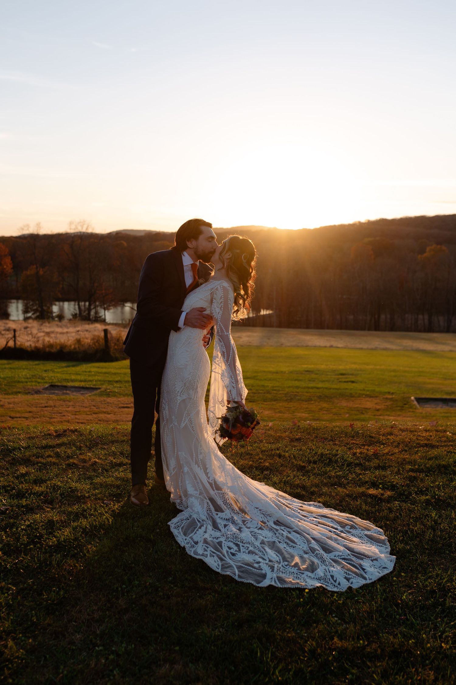 A couple in wedding attire embrace during golden hour sunset in a scenic countryside field with long lace dress trailing.