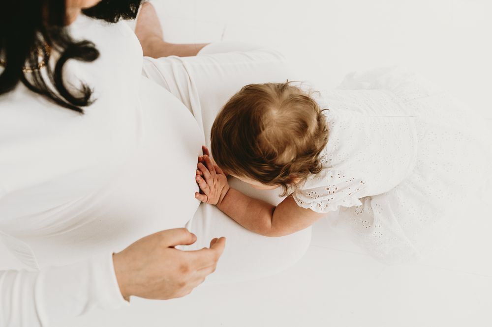Liten flicka håller på sin mammas gravidmage under en gravidfotografering i Jönköping / Mullsjö. Dom fotograferas i en vit studio.