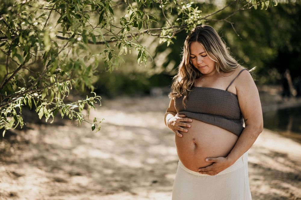 Gravid kvinna står på en strand i Jönköping.Hon tittar på sin bara mage. Det är sand och träd och vatten runt henne.