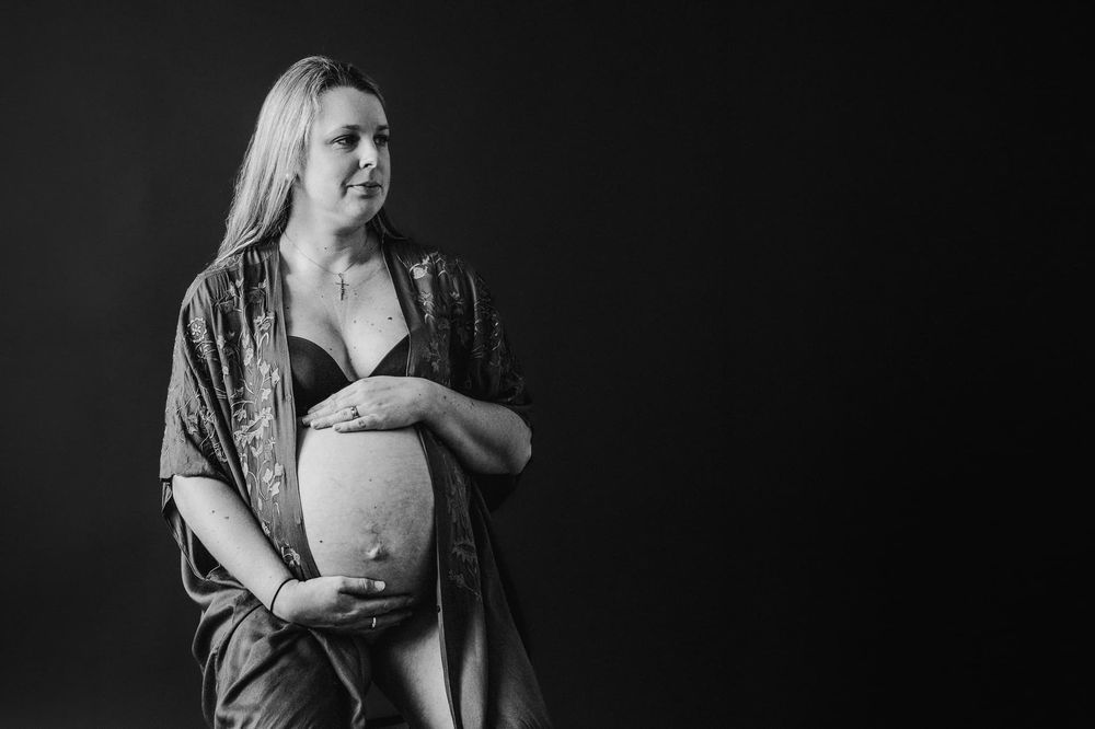 Gravidfotografering i studio. Gravidhåller om sin gravida mage. Hon har en kaftan och bh på sig, och en bar mage. Bilden är svartvit.