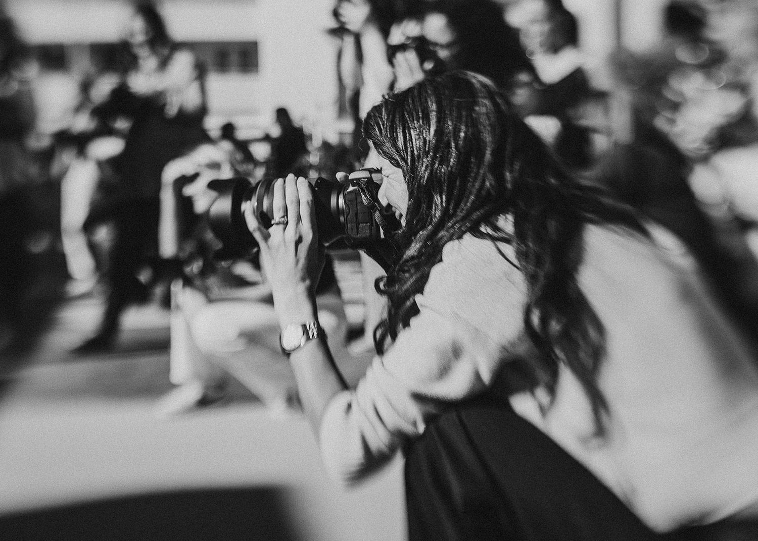 Black and white photograph showing photographer with long dark hair taking pictures in blurred background.