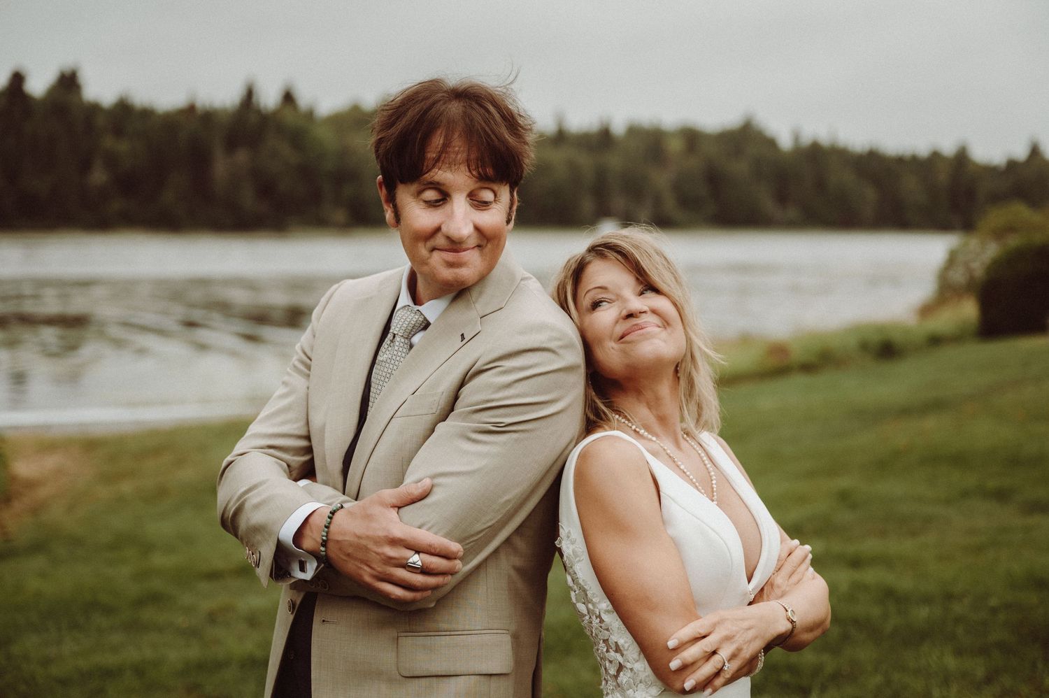 Couple poses back-to-back against lakeside backdrop during wedding photoshoot.