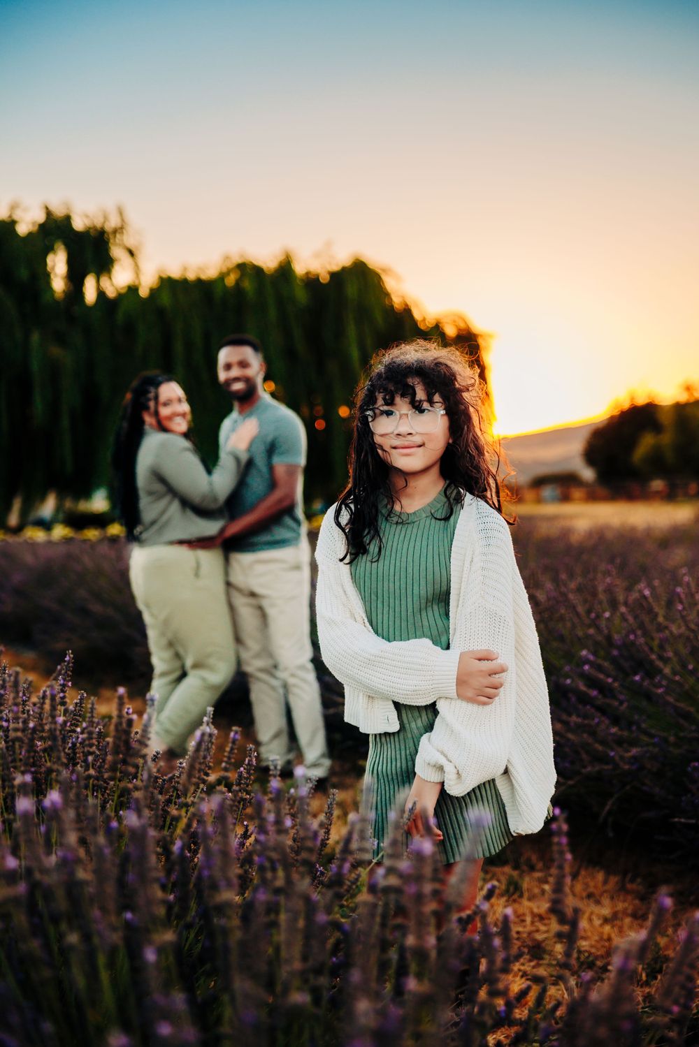 A family poses in a beautiful lavender field at sunset with warm golden light filtering through trees in background.