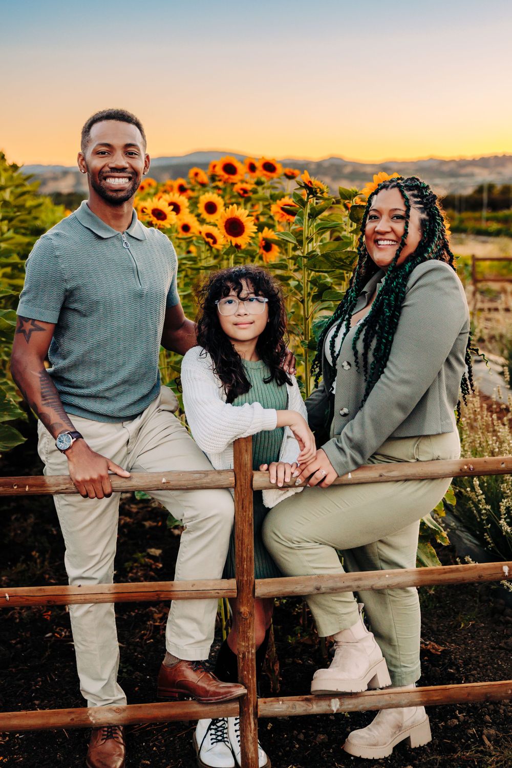 A loving family poses together on a wooden fence at sunset in front of vibrant blooming sunflowers.