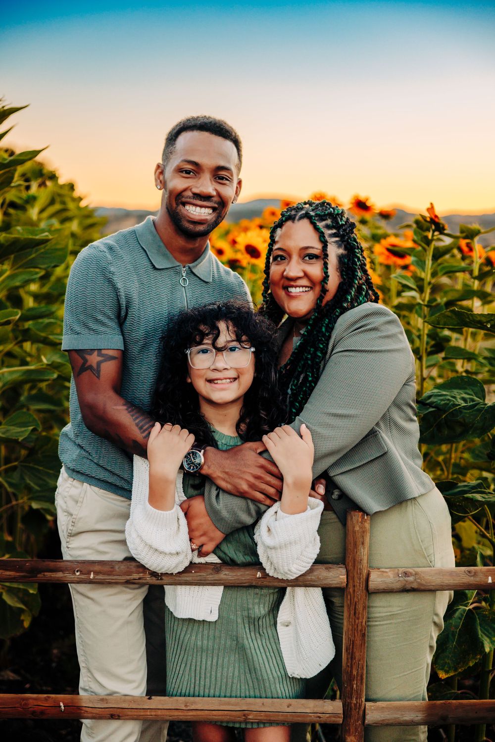 Family shares warm embrace while posing on rustic fence with golden sunflowers and sunset backdrop.