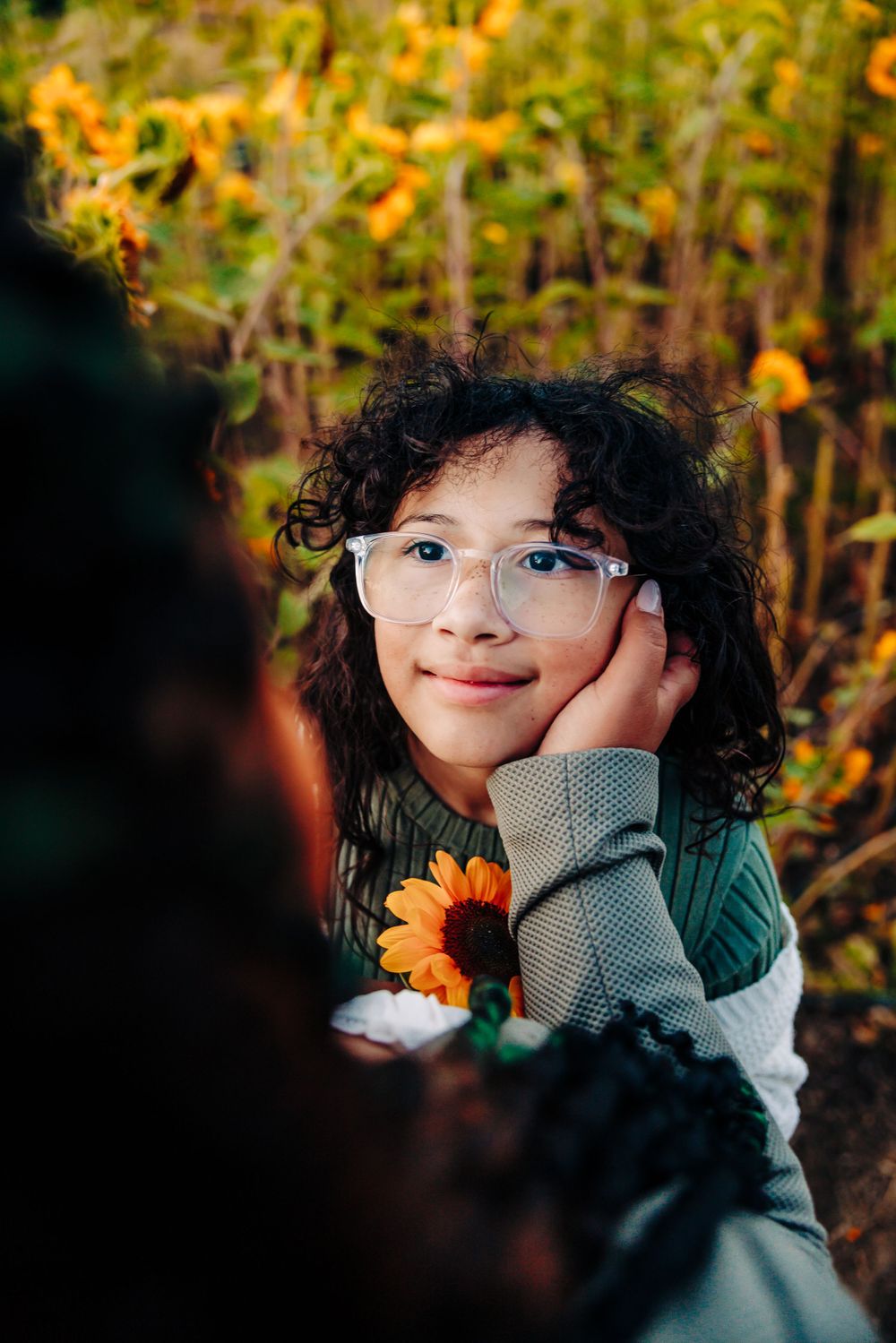 Portrait in autumn sunflower field with glasses and gray sweater showing gentle expression against golden background.