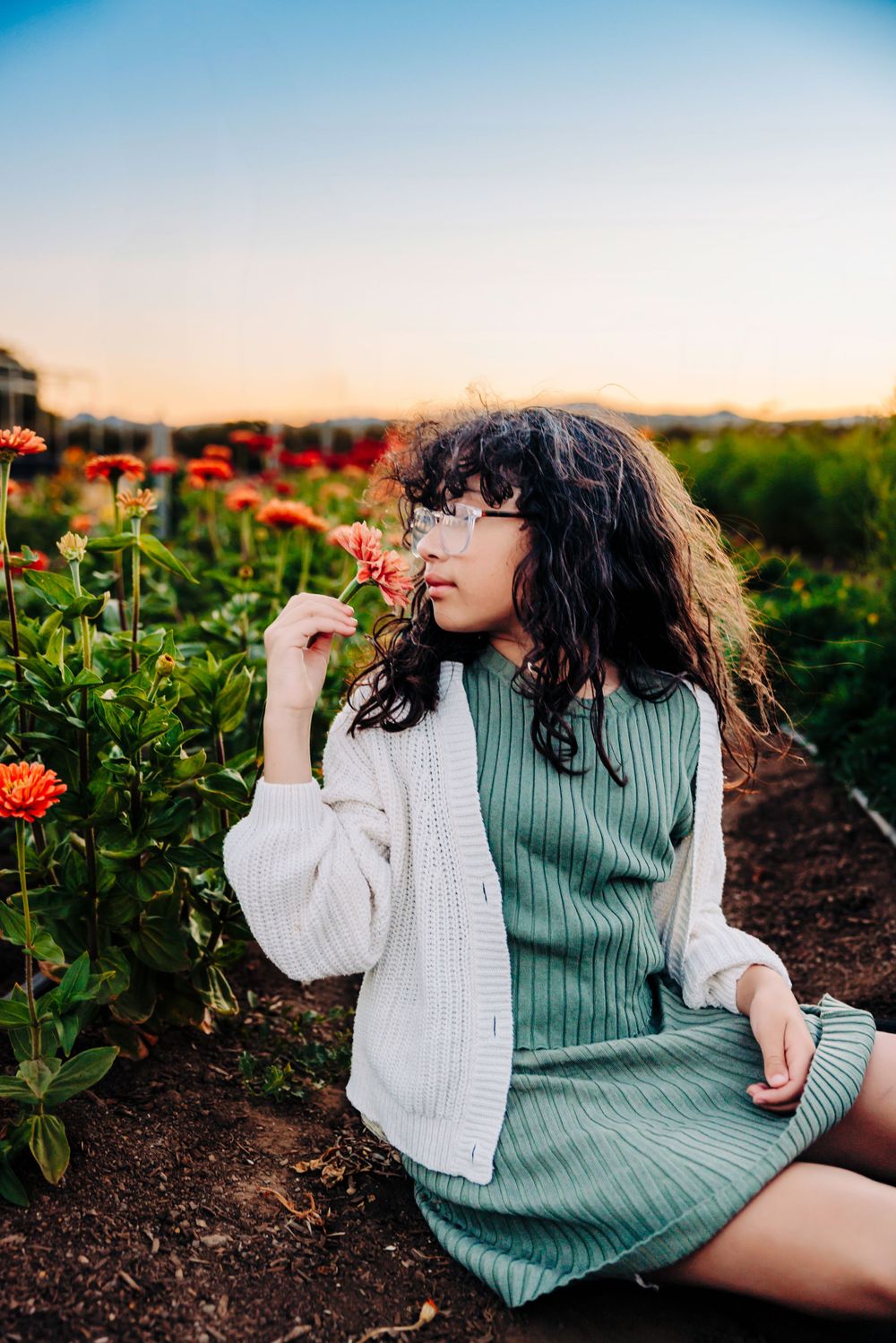 A person in a green dress and white cardigan poses among blooming orange dahlias at sunset in a garden.