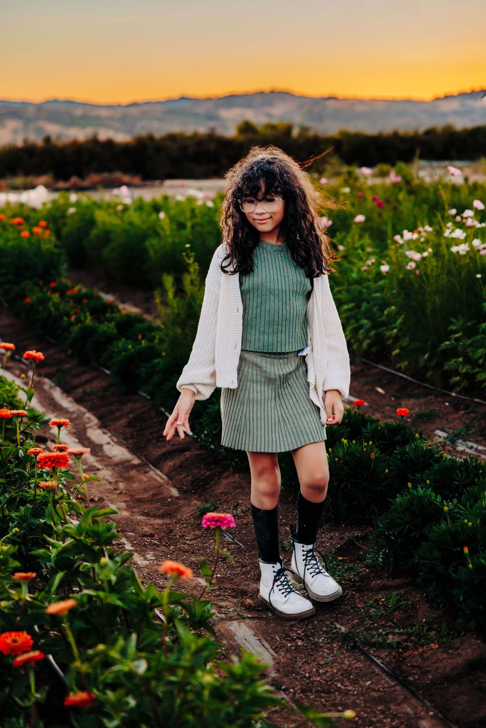 A youth stands in a flower field at sunset wearing a green knit dress, white cardigan and combat boots.