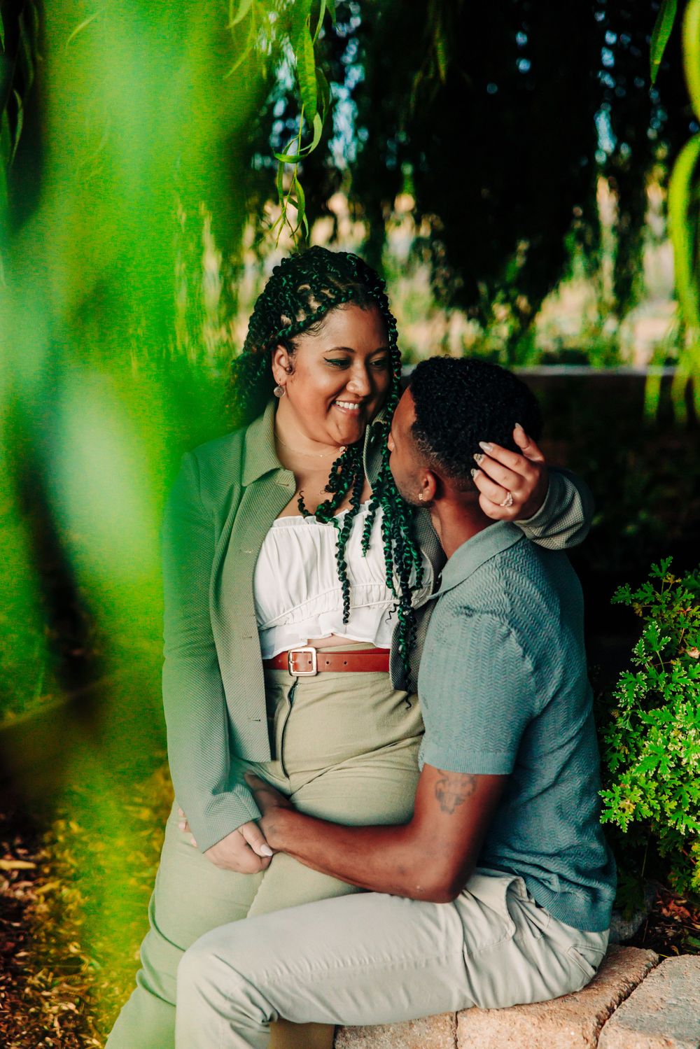A romantic couple shares a tender moment while sitting together under green foliage in a garden setting.