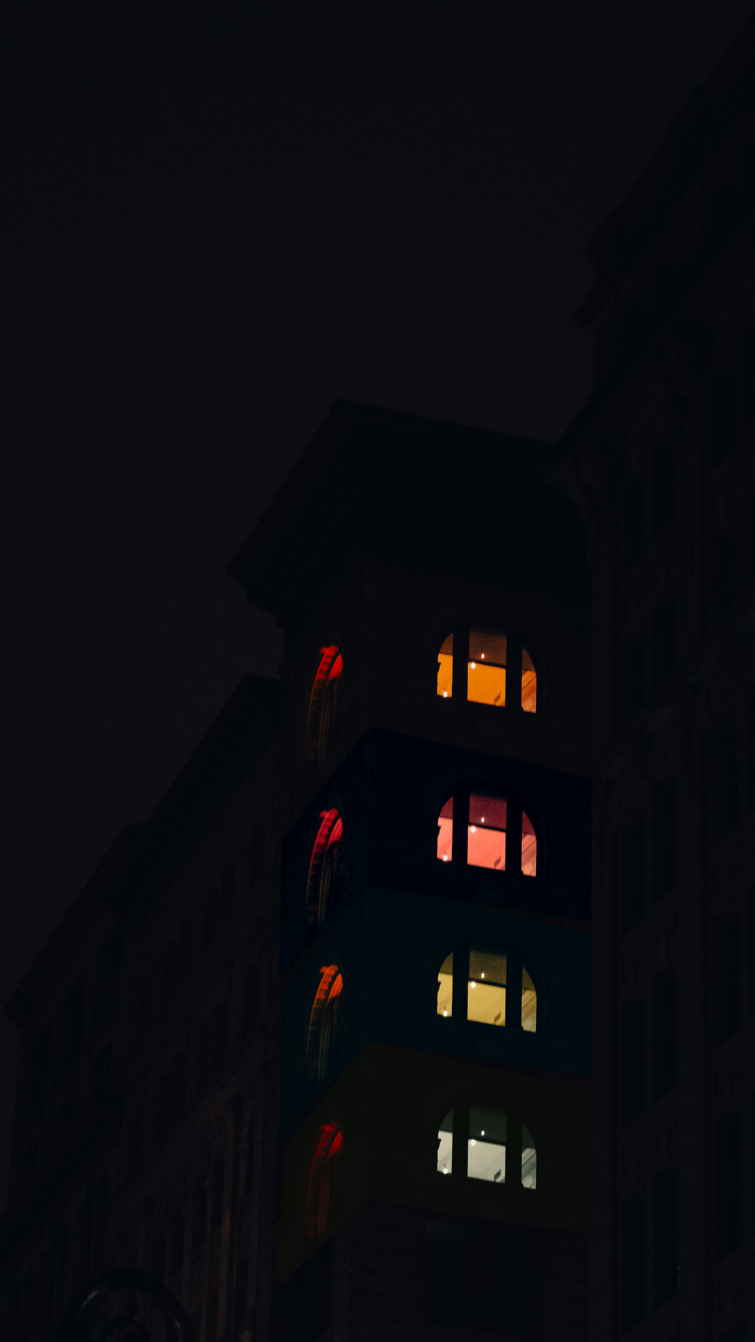 Tall dark building with illuminated windows glowing in warm orange and red colors against a black night sky.