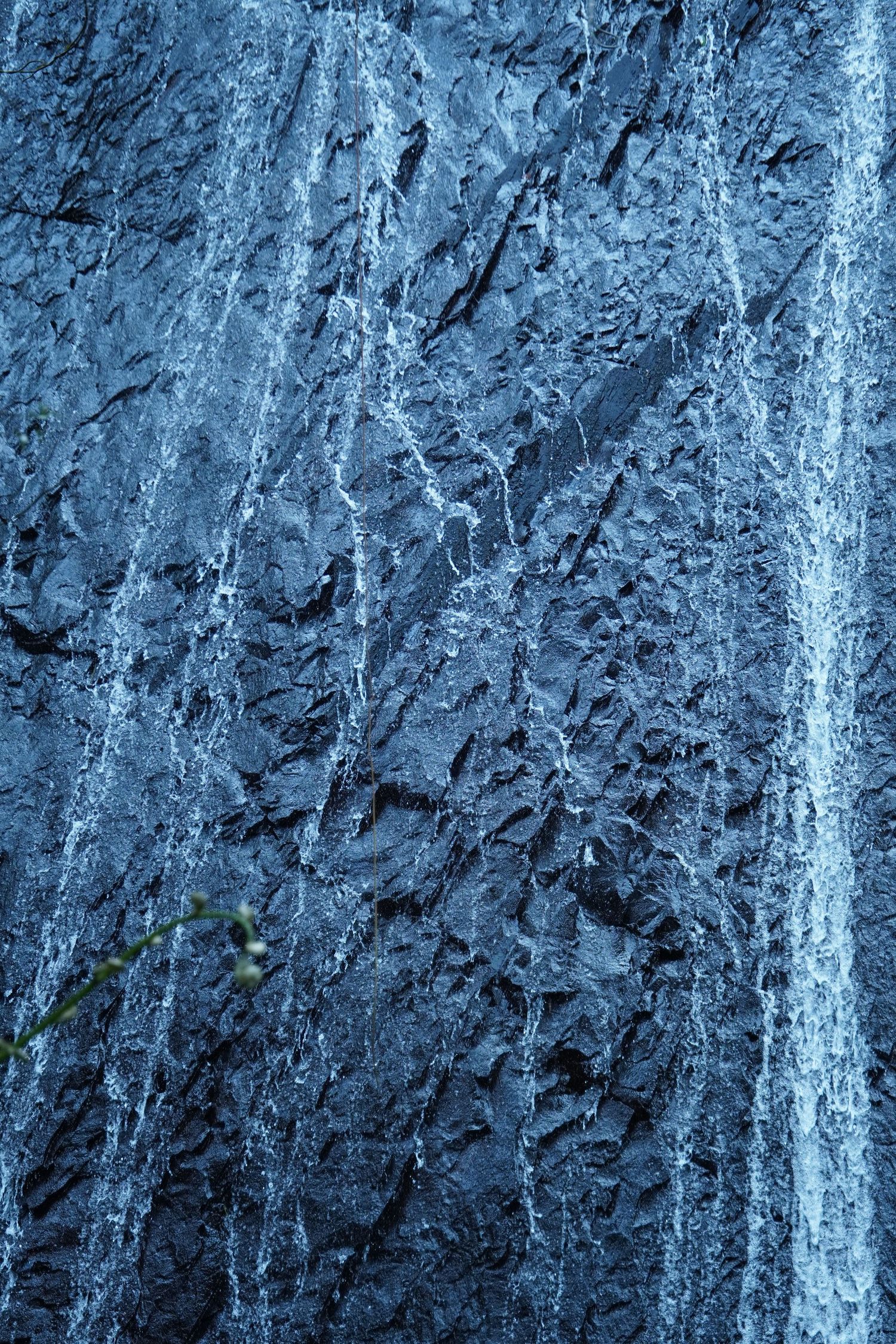 Textured dark stone wall with streaks of water running down its rough surface creating a moody blue atmosphere.