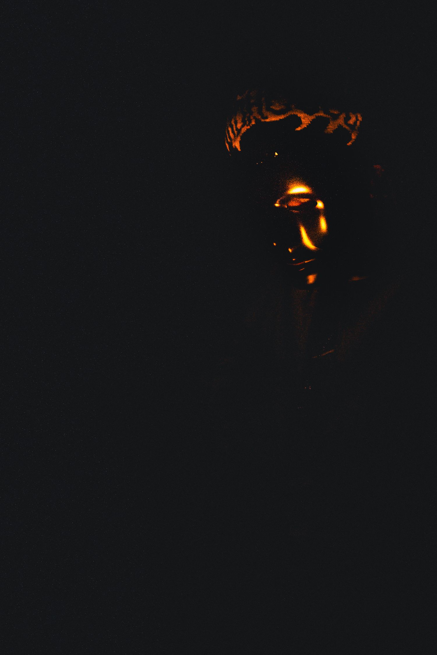 Dark moody portrait illuminated by warm orange lighting revealing subtle facial features in shadow.