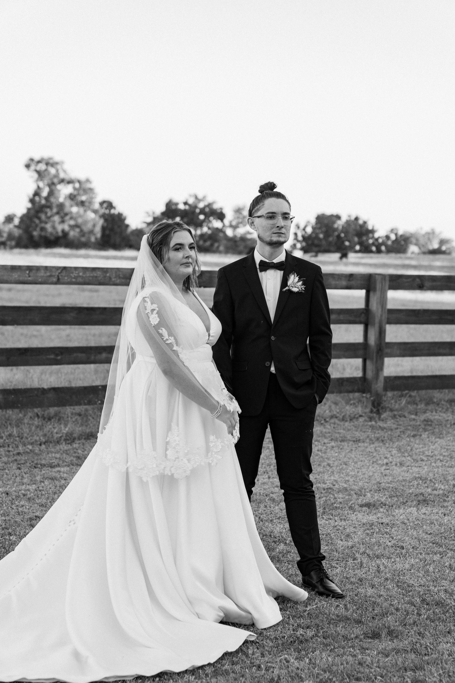 Black and white wedding photo of a bride and groom posing together in front of a rustic wooden fence.