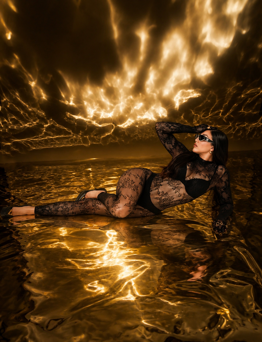 Woman in black lace bodysuit poses in golden water under dramatic fiery sky.