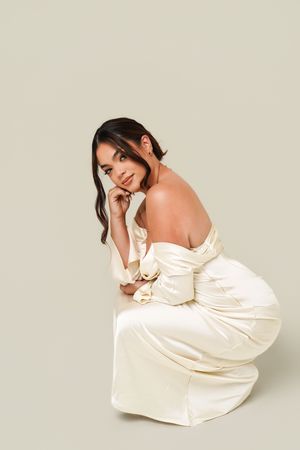 Woman in elegant ivory satin off-shoulder gown crouching gracefully against neutral background.