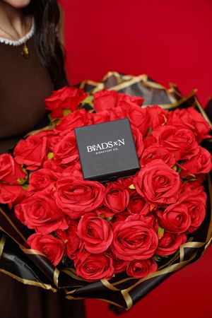 Elegant arrangement of vibrant red roses with a black Bradsons gift box against a dark background.