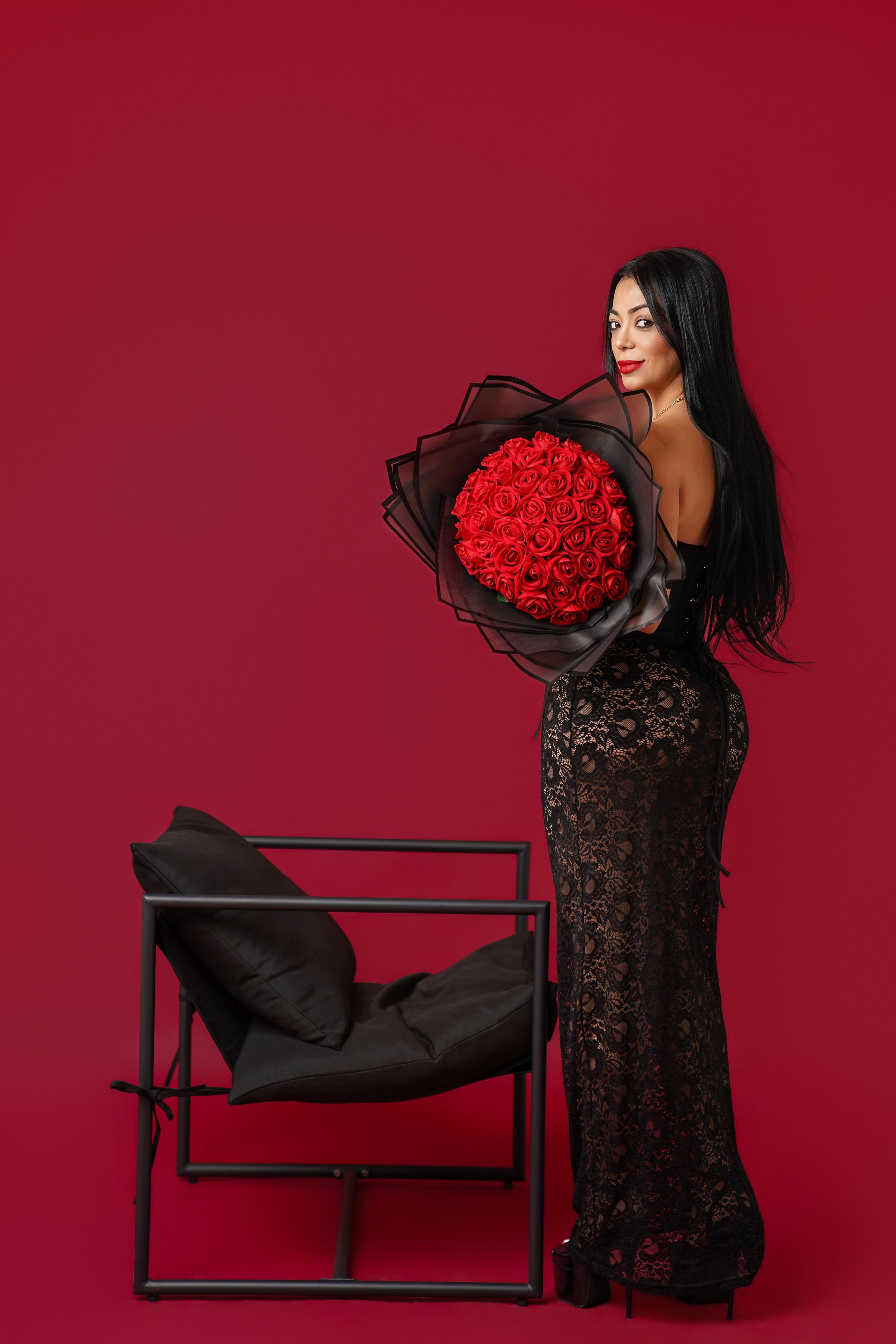 Elegant woman in black lace gown holding large red rose bouquet, posing by modern chair on red background.
