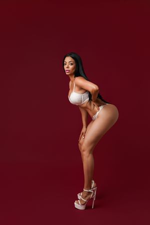 Woman in white lingerie and heels posing confidently against a deep red background.