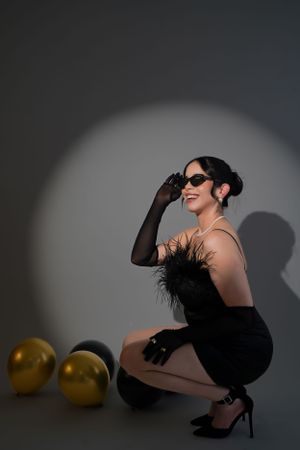 Fashion model in black attire and sunglasses poses artistically with golden balloons against dark backdrop.