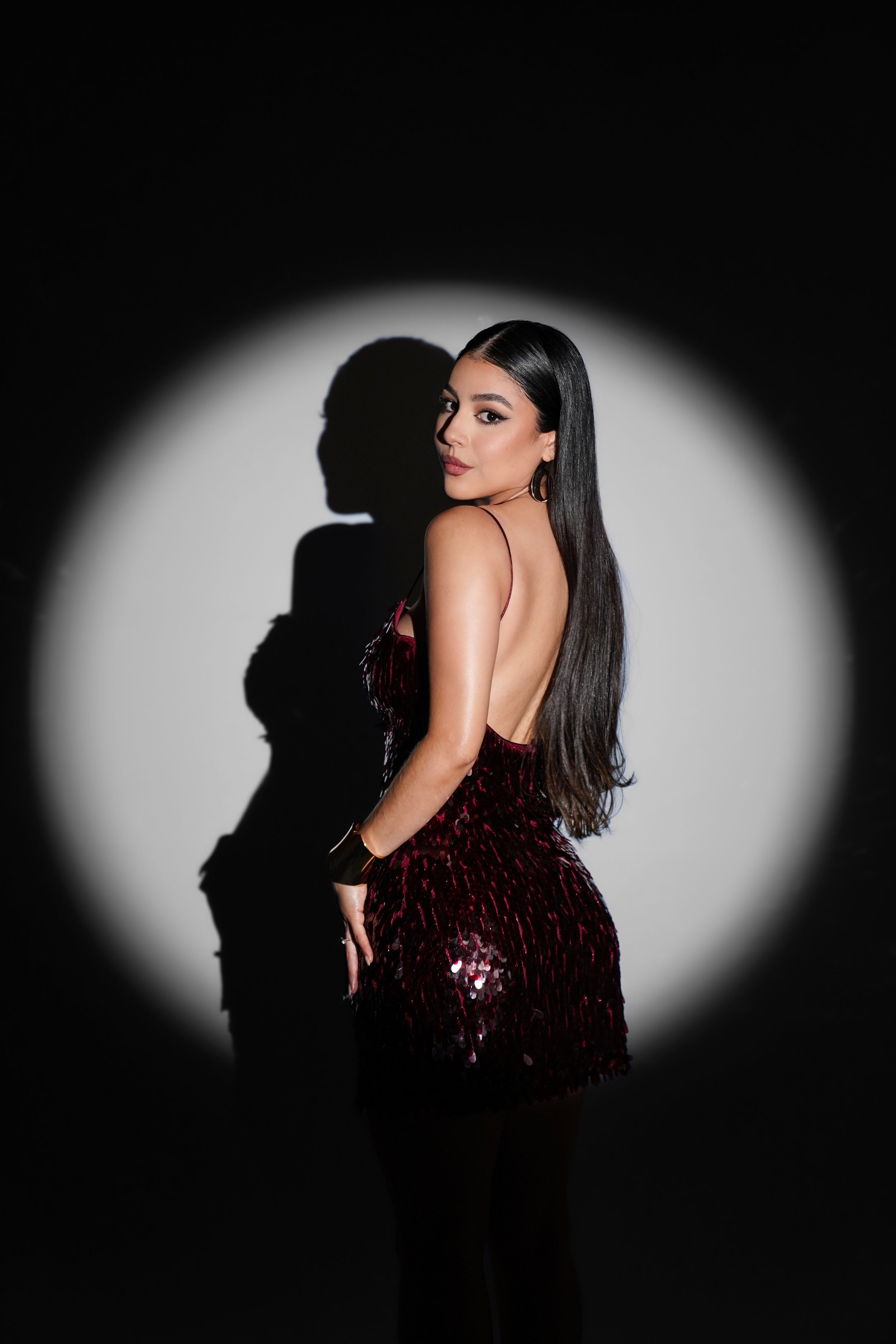 Woman in burgundy sequin dress poses against dark background with dramatic shadow and spotlight effect.