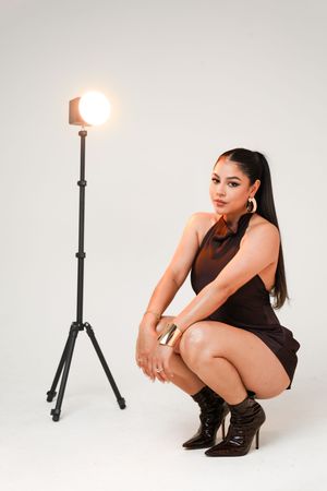 Studio portrait with a figure in brown dress crouching next to a lighting stand against white backdrop.