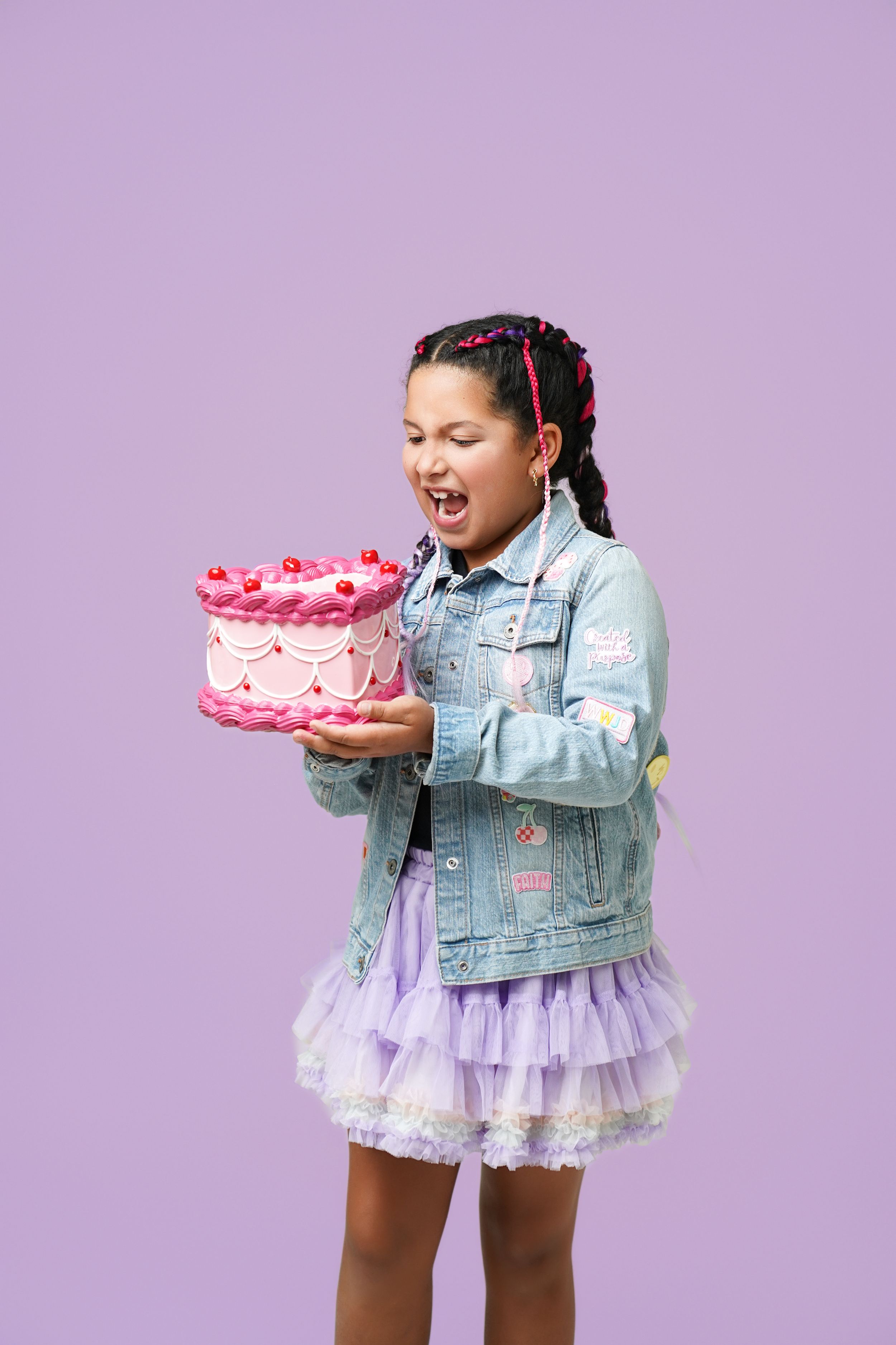 Excited young girl in denim jacket holding a pink birthday cake against a purple background.