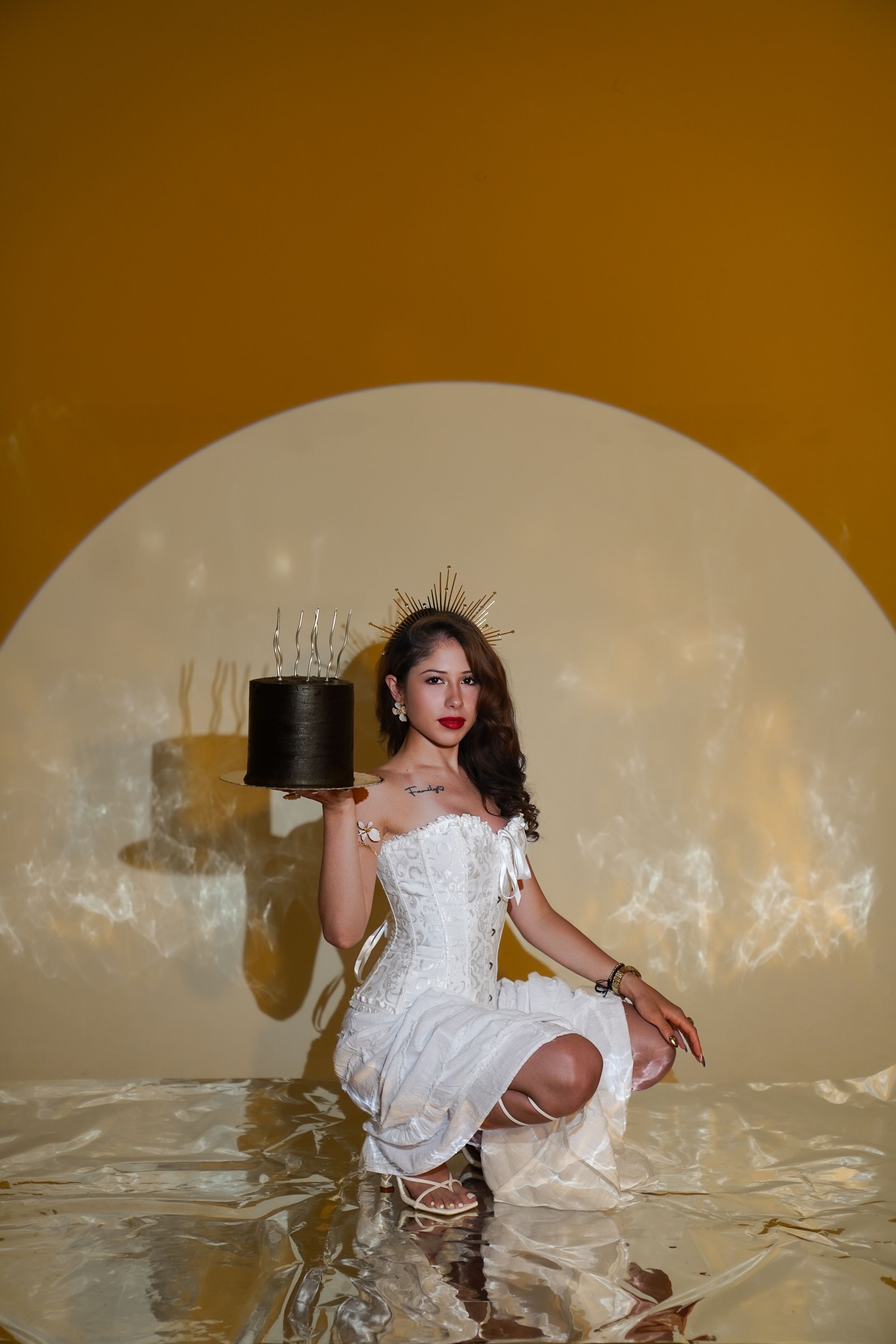 Woman in white corset dress holding black cake with crown, posing against golden yellow background with circular light.
