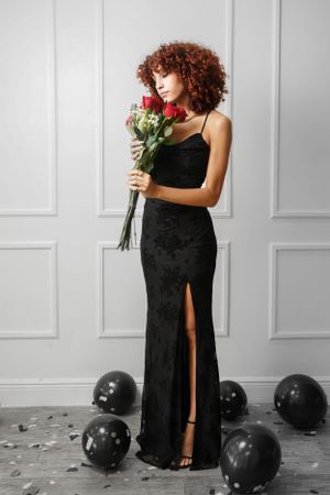 Person in elegant black dress holds red roses against white paneled wall with black balloons.
