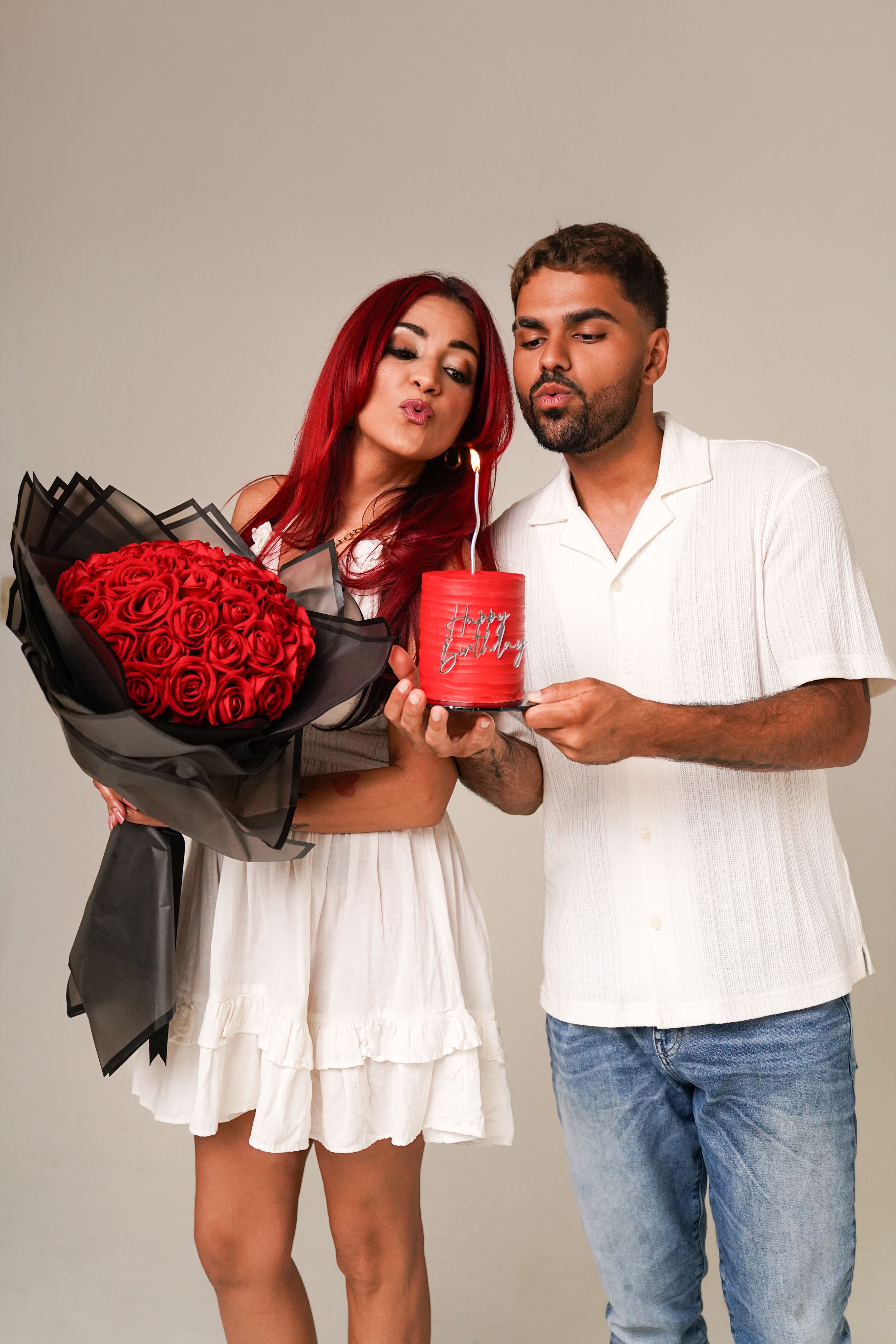 Couple in white outfits blowing on a red gift box, woman holding large red rose bouquet wrapped in black paper.