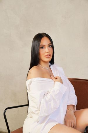 Woman in white loungewear poses on brown leather couch in studio setting.