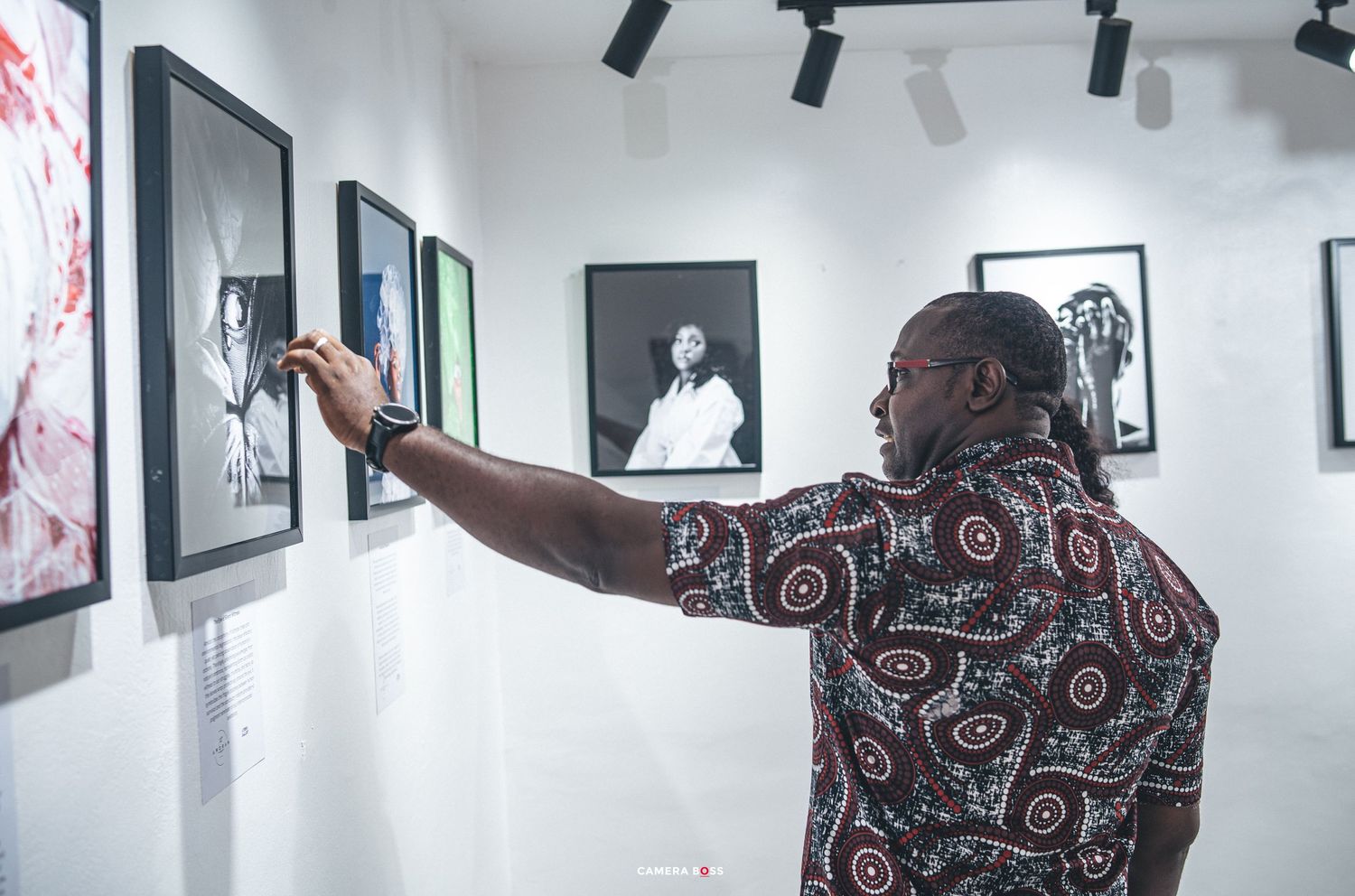 Person in patterned shirt examines artwork displayed in gallery with track lighting and white walls.