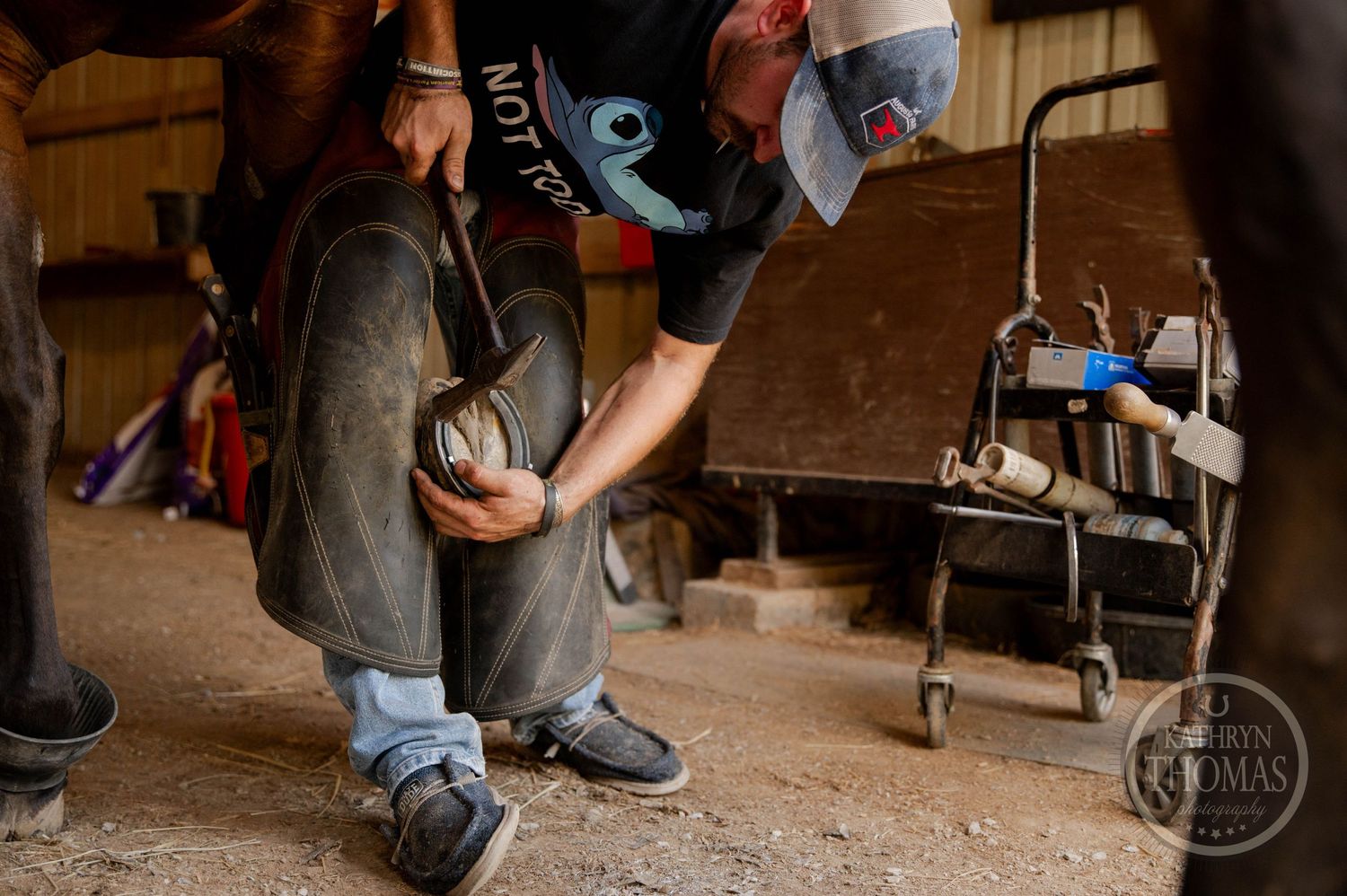 A farrier kneeling to work on horseshoes in a dimly lit stable.