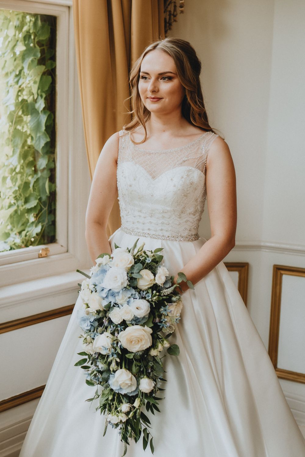 Bride in elegant white gown with illusion neckline holds cascading bouquet of white roses by sunny window with ivy.