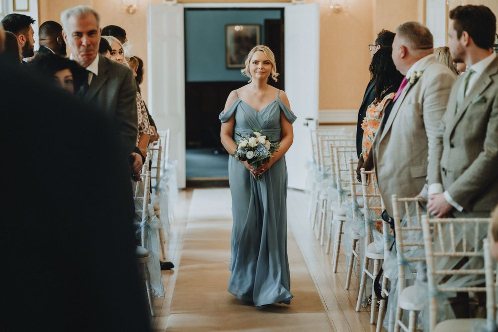 A wedding party member in a flowing blue gown walks down the aisle during a ceremony surrounded by guests.