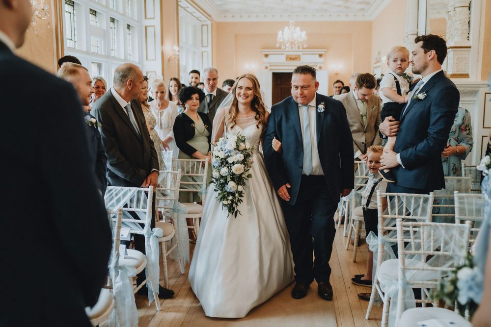 An elegant wedding ceremony processional in a bright room at Holmewood Hall with white chairs and decorative floral arrangements.