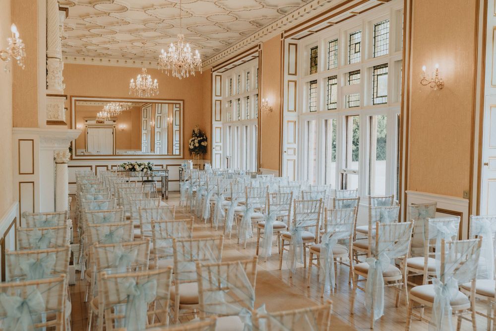 Elegant wedding ceremony at Holmewood Hall with white chairs and pink walls illuminated by crystal chandeliers and natural light.