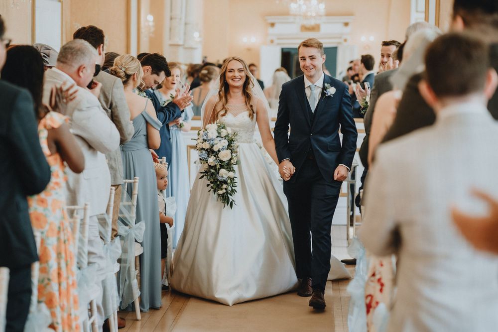 A bride and groom walk down the church aisle lined with guests, as natural light fills the ceremony space.