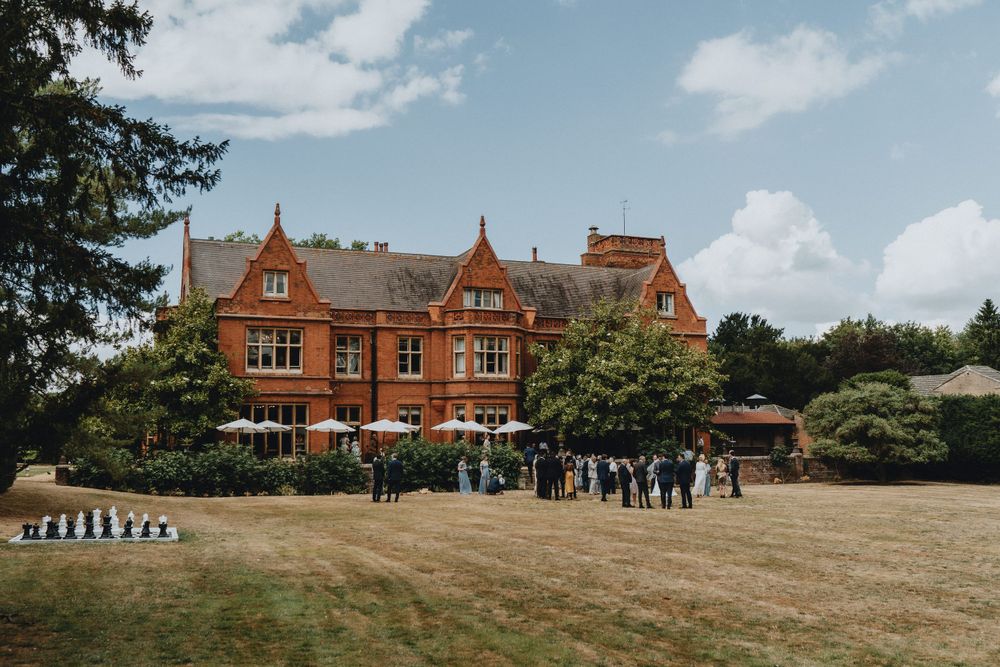 A large Victorian red brick mansion of Holmewood Hall with guests gathered outside on the lawn on a sunny summer day.