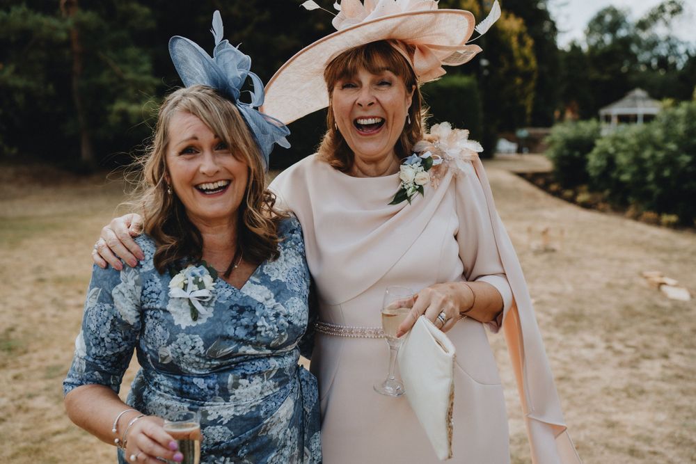 Wedding guests in elegant floral dresses and fancy hats sharing a joyful moment at an outdoor celebration.