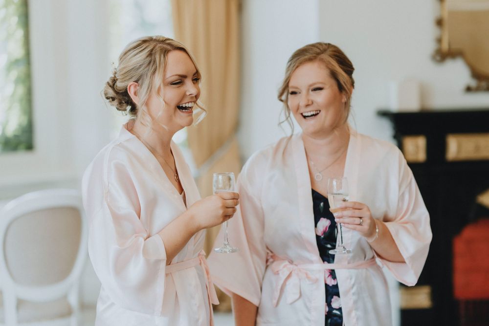 Two women in matching pink robes share a laugh while holding drinks during a wedding preparation moment.