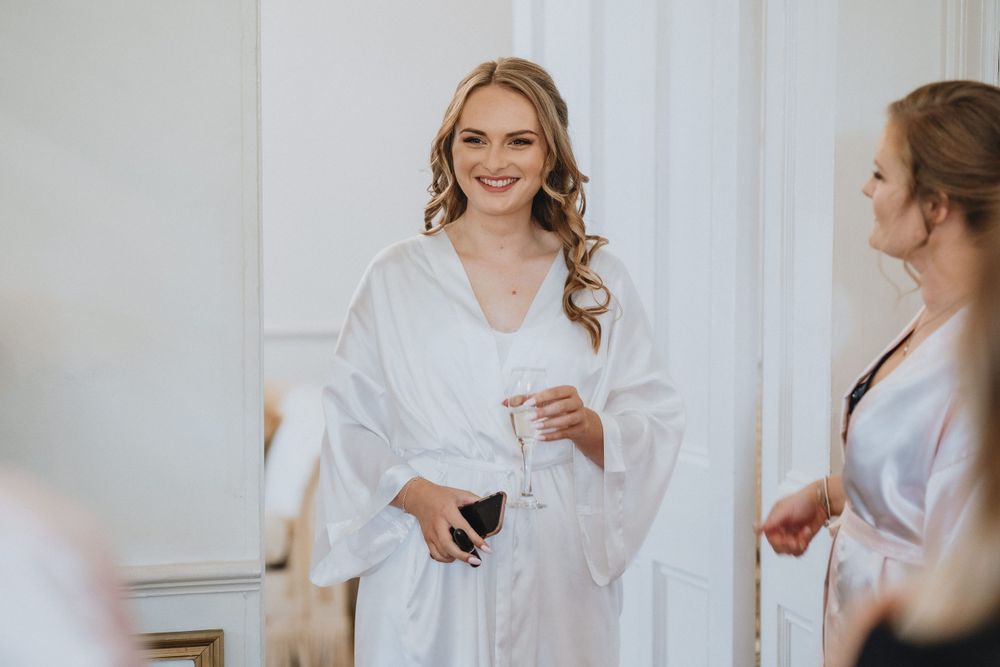 Woman in white silk robe smiles while getting ready in elegant dressing room with mirror.