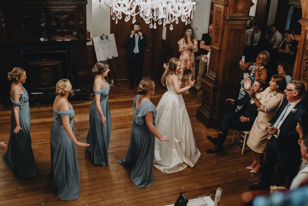 Wedding party stands in formation on wooden dance floor under elegant chandelier during formal ceremony.