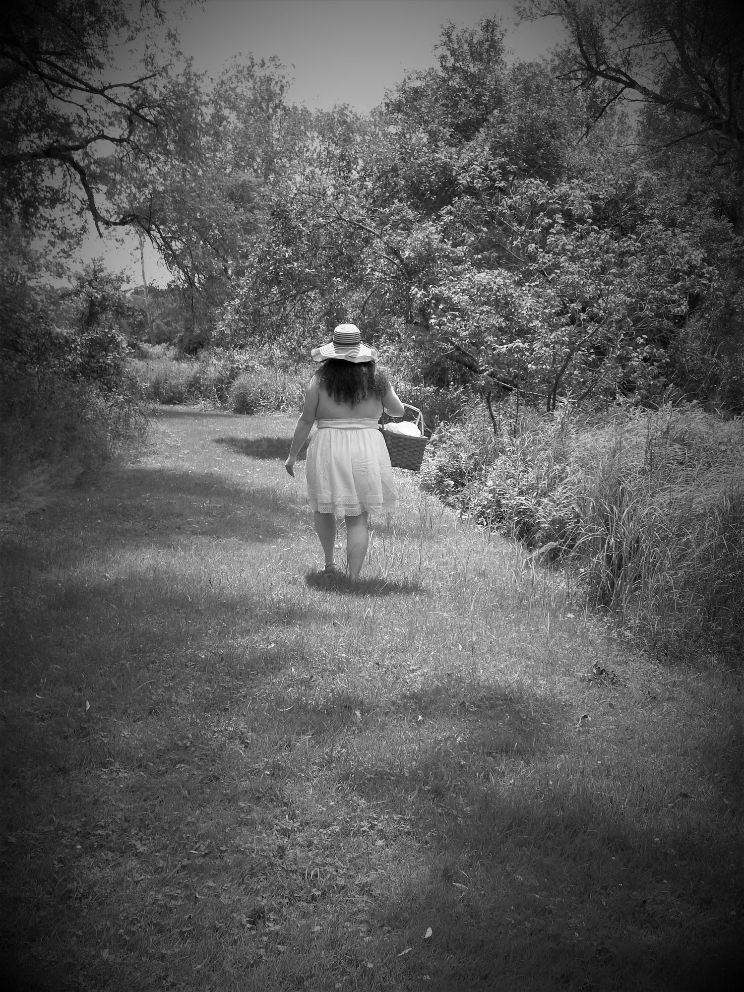 Black and white image of someone walking through a grassy field wearing a sun hat.