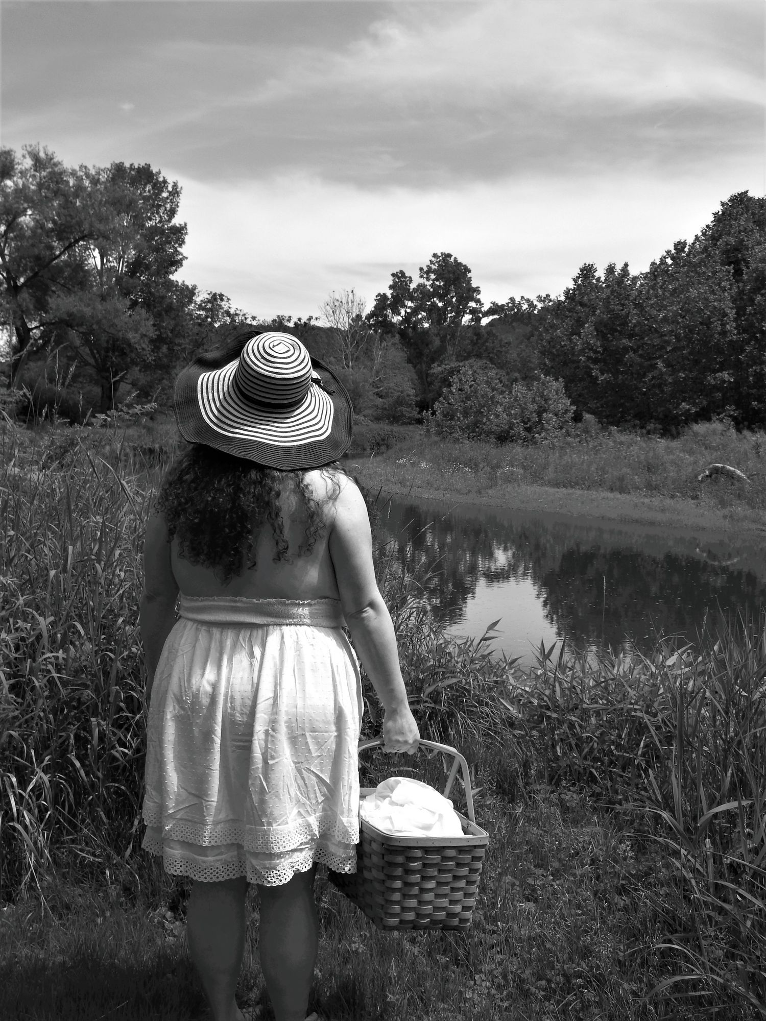 Figure in white dress and sun hat walking along a pond carrying a woven basket.