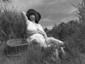 A black and white photo of someone reclining in a white dress and striped sunhat amid tall grass near a lake.