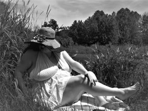 Artistic black and white photograph of someone lounging in nature wearing a sunhat and white dress.