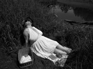Black and white artistic photograph of someone reclining in a white dress by a lake with dramatic lighting.