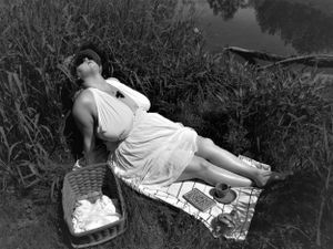 Monochrome image of a figure in white lying near water's edge with natural foliage in background.