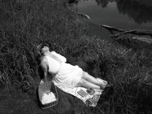 Artistic black and white landscape photo with person in flowing white dress resting by water.